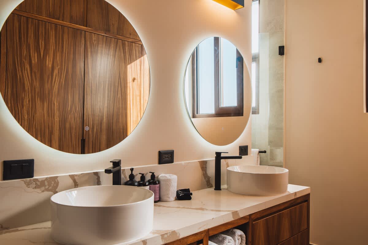 A contemporary double vanity bathroom showcases two white circular vessel sinks set on a marble-patterned countertop, flanked by two large round mirrors with integrated backlighting. Dark wooden cabinetry below and sleek black faucets complete the sophisticated look.
