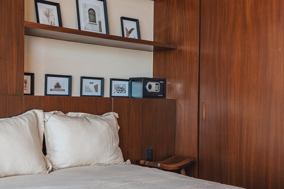 Cozy bedroom featuring a bed with white bedding, a dark wooden headboard with integrated shelving displaying framed art, and a built-in wooden wardrobe with a safe.