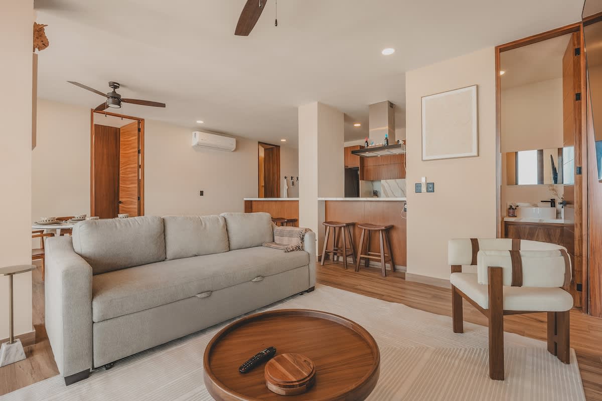 Comfortable living room featuring a large light gray sofa, a round wooden coffee table, and a wall-mounted TV, with an open view to the kitchen and dining area.