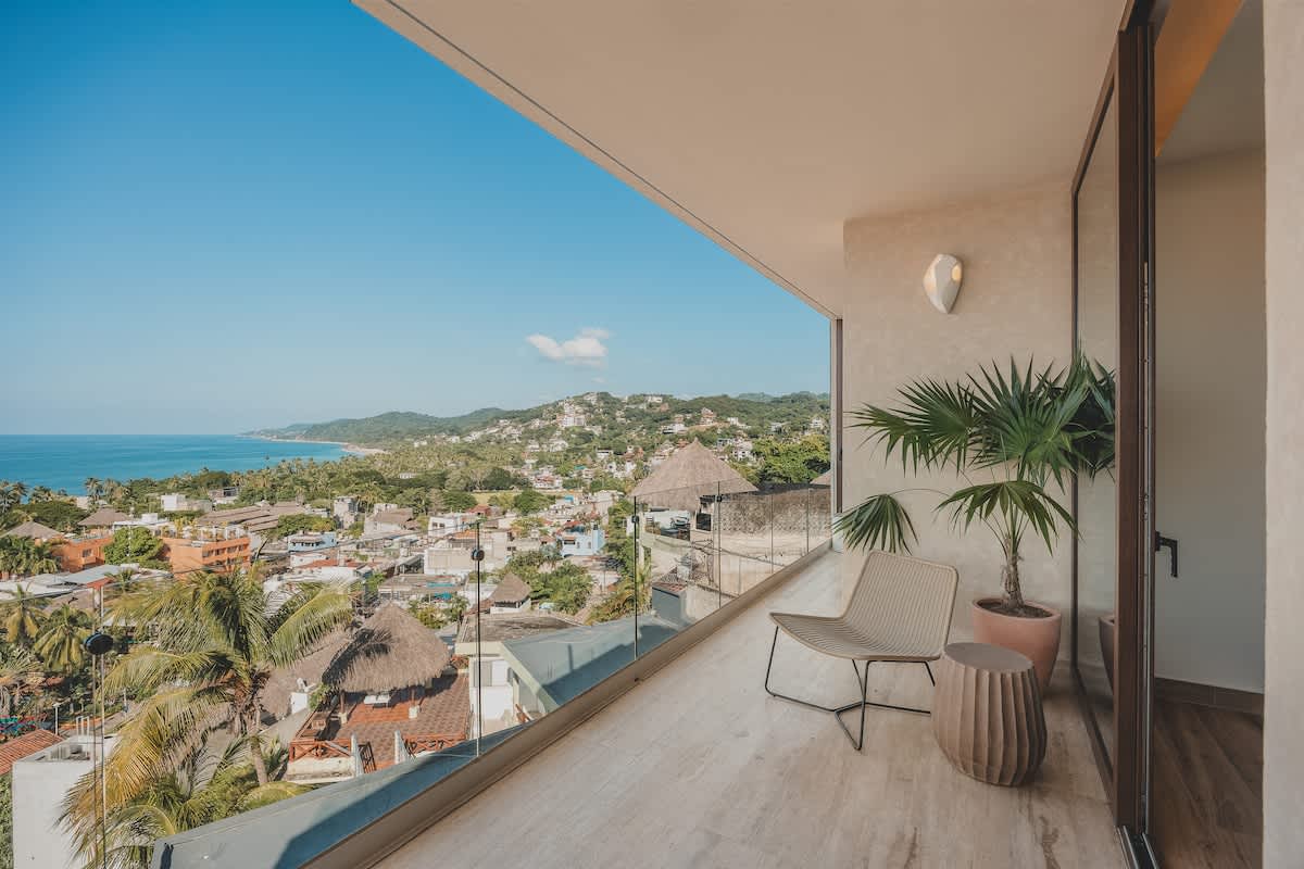 Spacious balcony offering panoramic views of the ocean and a vibrant coastal town, furnished with a modern lounge chair and a small side table, surrounded by a clear glass railing.