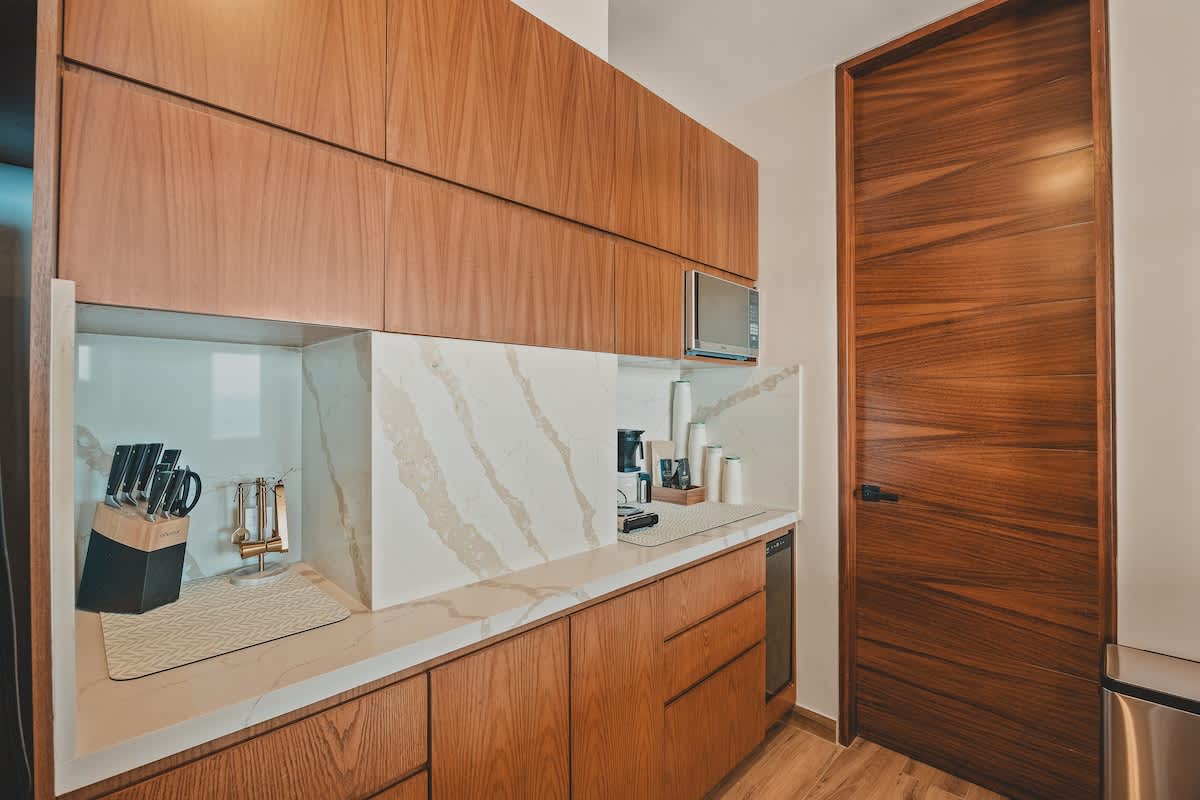 Close-up of a section of the modern kitchen with warm wooden upper and lower cabinets, white marble countertops and backsplash, and a built-in microwave, featuring a knife block and coffee maker.