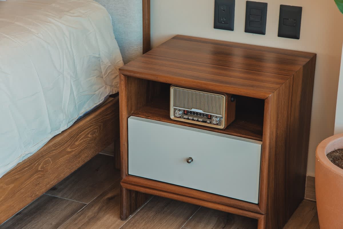 Detail shot of a modern wooden nightstand with a light gray drawer and a vintage-style radio, positioned next to a bed with white bedding.