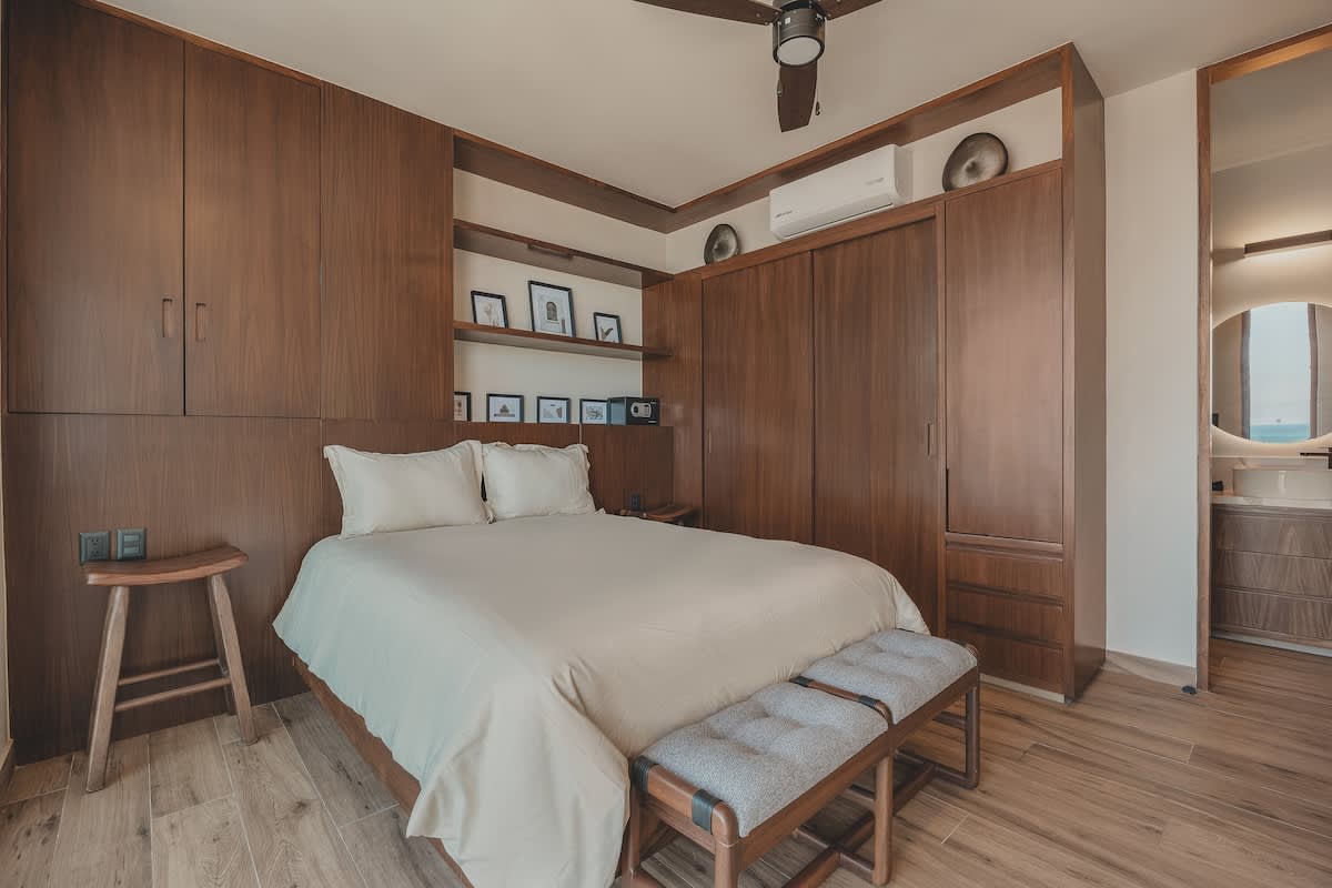 Master bedroom featuring a queen bed with two upholstered benches, extensive built-in wooden wardrobes and shelving, a ceiling fan, and a glimpse of the en-suite bathroom.