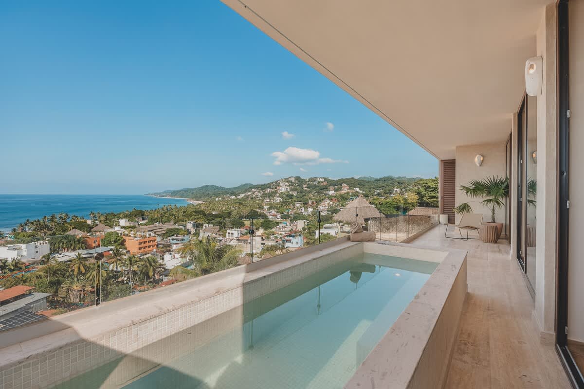 Panoramic view from a spacious balcony showcasing a private plunge pool and the vast expanse of the ocean meeting the horizon, with a vibrant coastal town nestled below.
