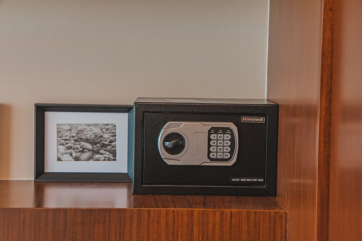 A black Honeywell digital safe with a silver keypad is placed on a polished wooden shelf, next to a framed black and white photograph. The safe is compact and modern, blending with the neutral wall behind.