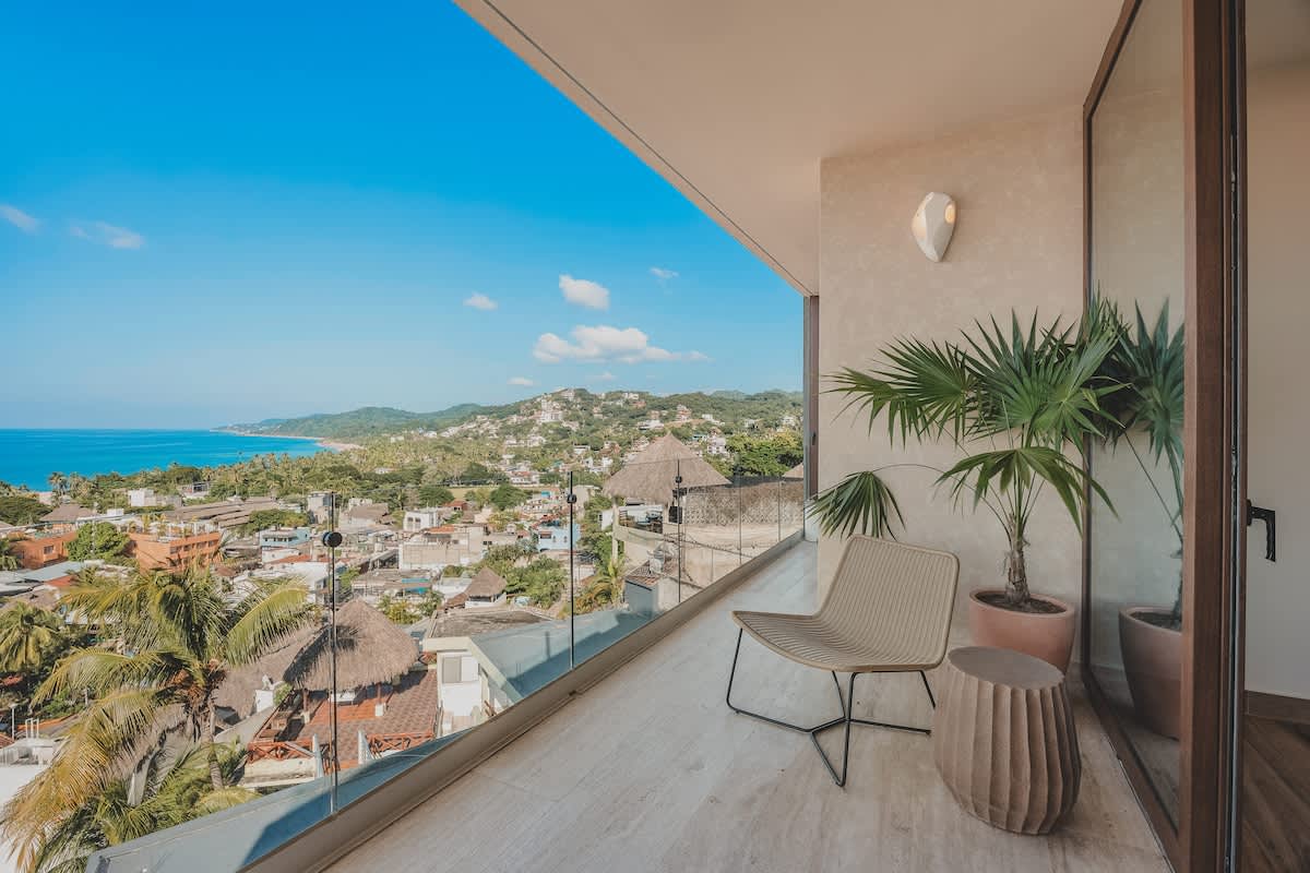 Spacious balcony with a modern lounge chair and a side table, offering expansive panoramic views of the ocean, coastline, and a vibrant town below.