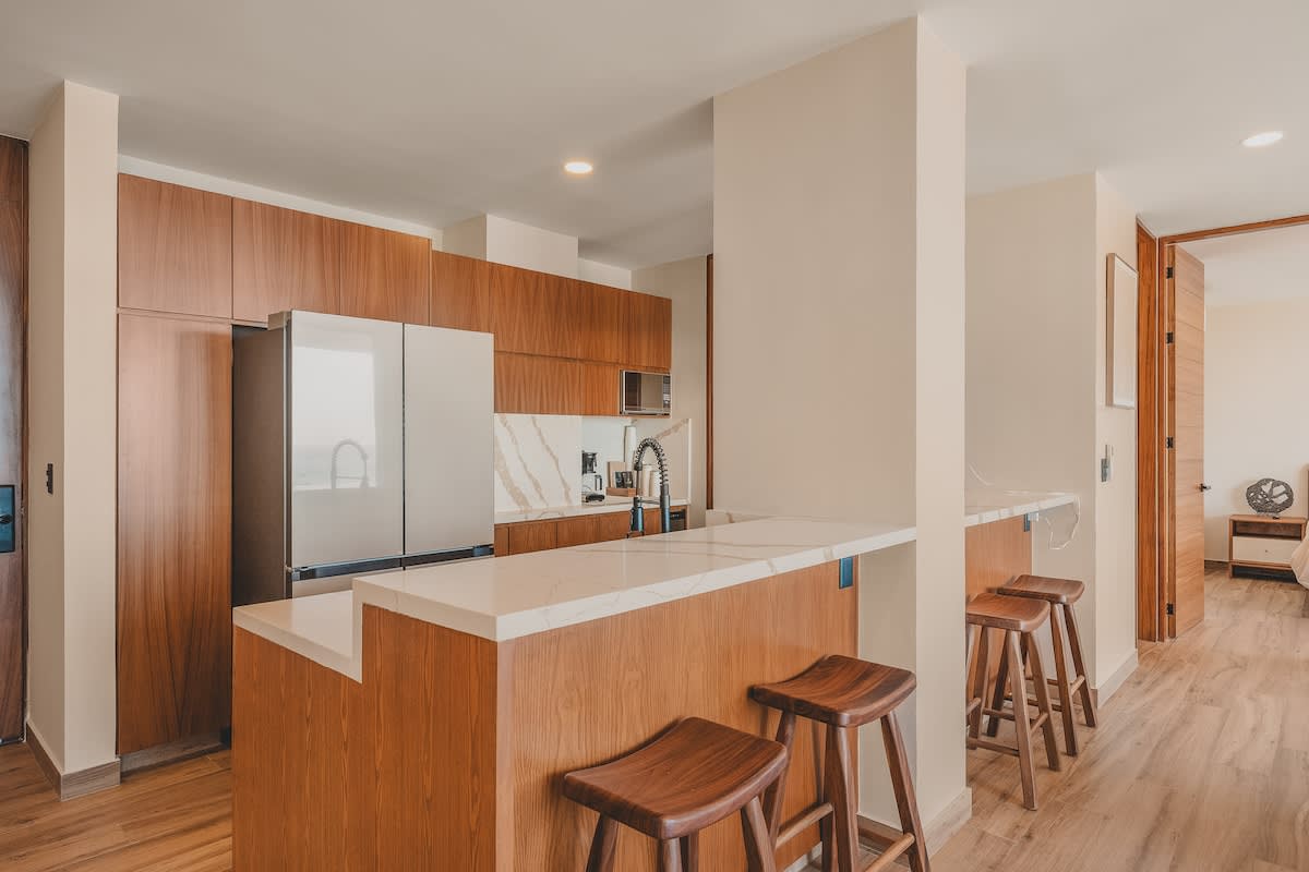 Modern kitchen featuring warm wooden cabinetry, white marble countertops and backsplash, and stainless steel appliances, with a breakfast bar and three wooden stools.