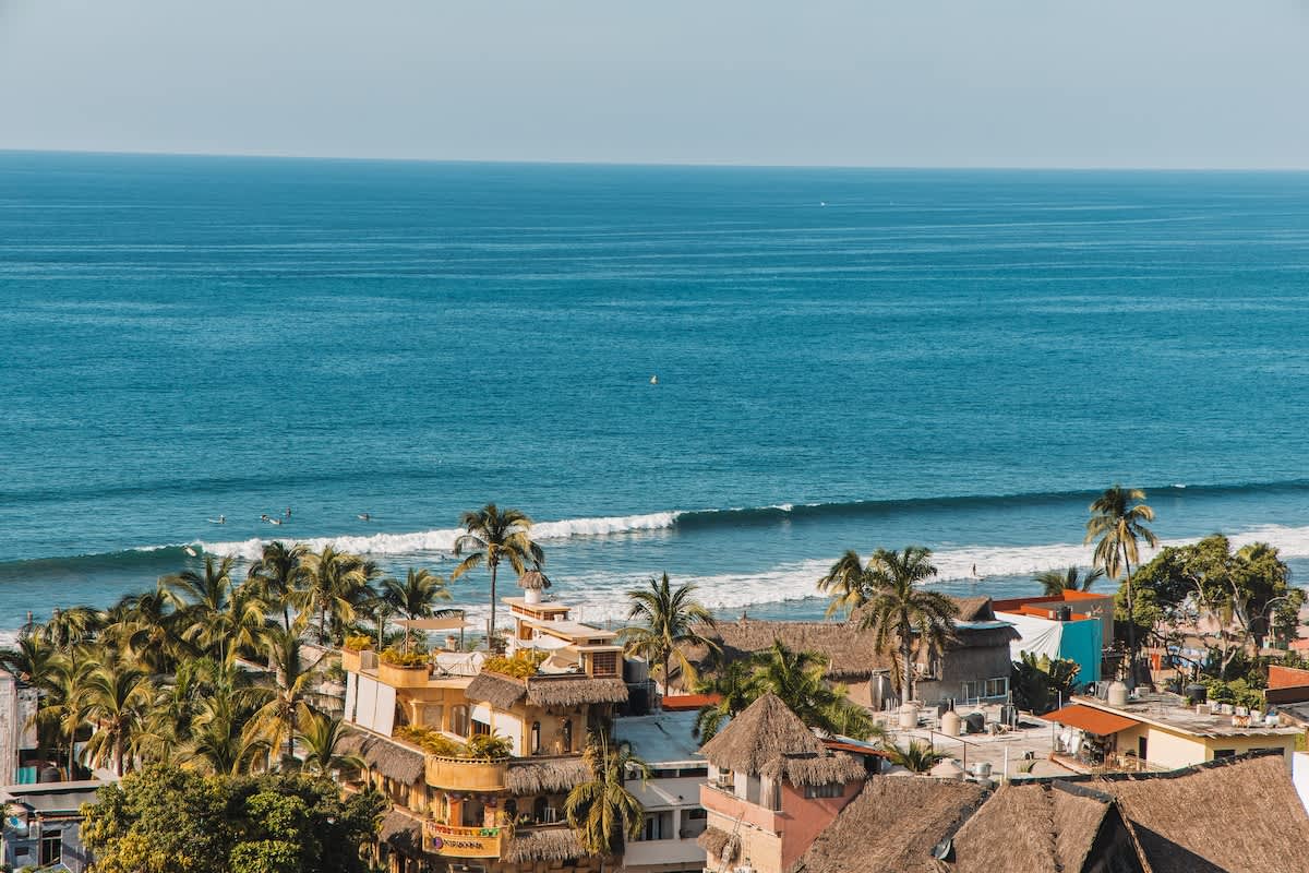 Aerial view of a coastal town with buildings featuring thatched roofs and palm trees, situated along a sandy beach with waves breaking in the clear blue ocean.