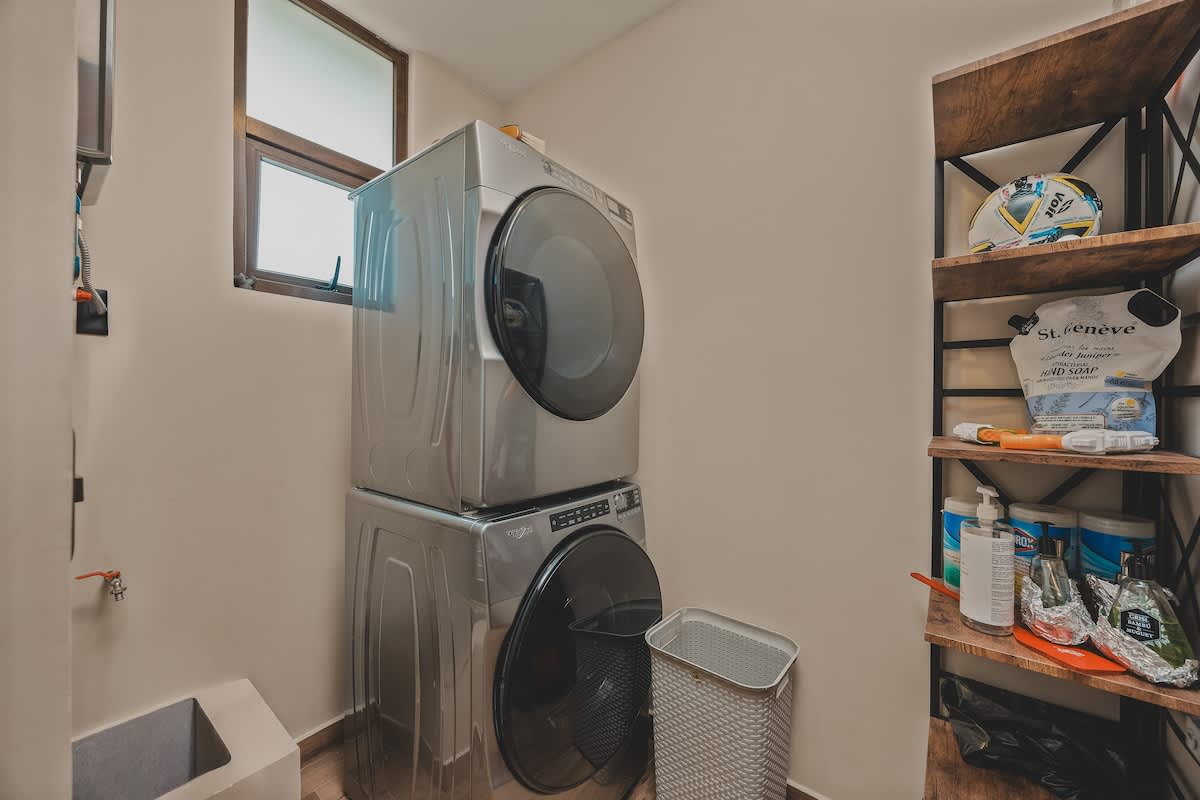 A functional laundry room features a stacked silver washer and dryer unit, positioned beneath a small window for natural light. A utility sink is visible on the left, while a rustic wooden shelving unit on the right holds cleaning supplies and other household items.