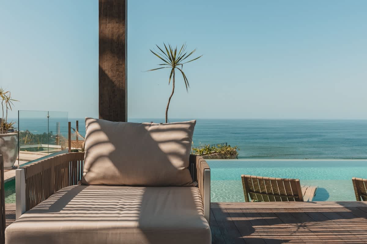 Comfortable outdoor chaise lounge with a beige cushion, positioned on a wooden deck beside an infinity pool, offering serene ocean views under a clear, sunny sky.