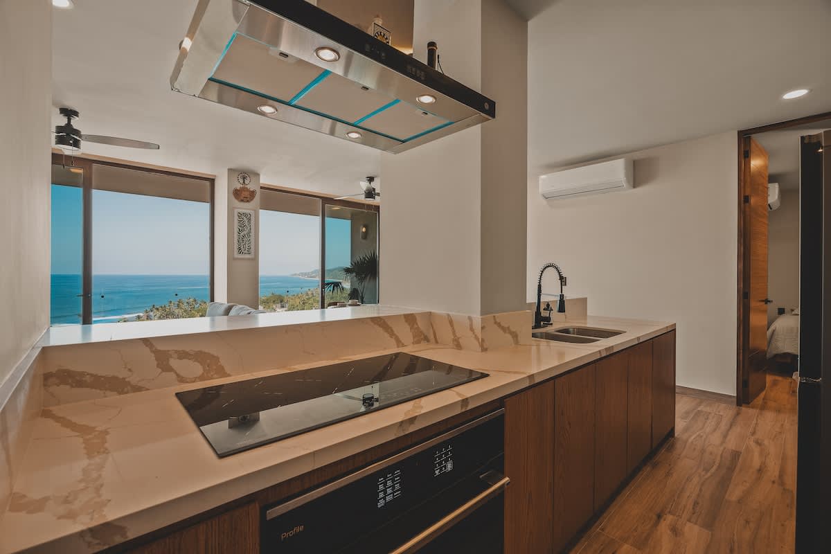 Modern kitchen with an induction cooktop and oven, featuring warm wooden cabinetry, white marble countertops, and a stainless steel range hood, with a glimpse of the ocean view through large windows.