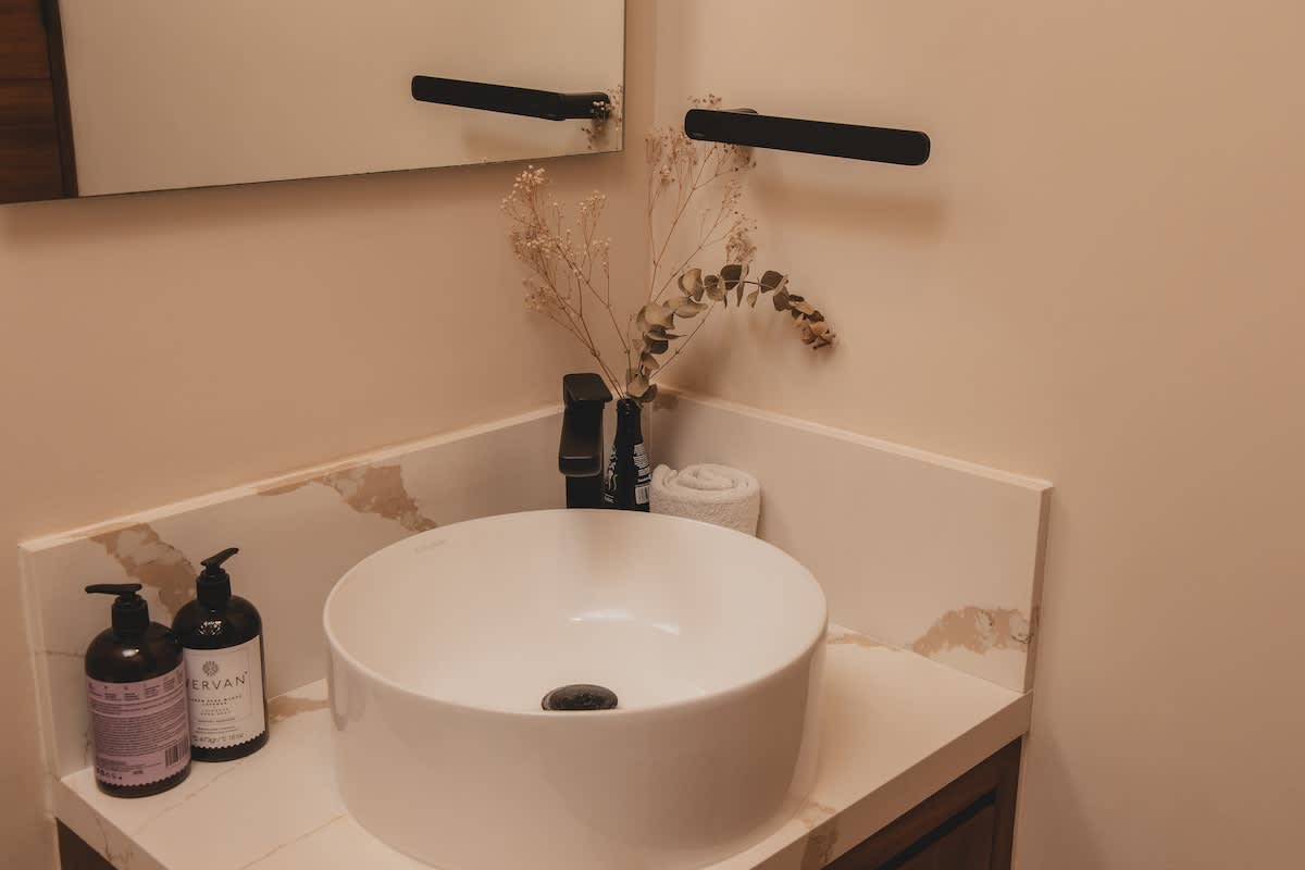 A modern corner bathroom features a white circular vessel sink on a light-colored quartz countertop with subtle brown veining. Black fixtures, including a faucet and two soap dispensers, complement the minimalist design, alongside a small dried floral arrangement.