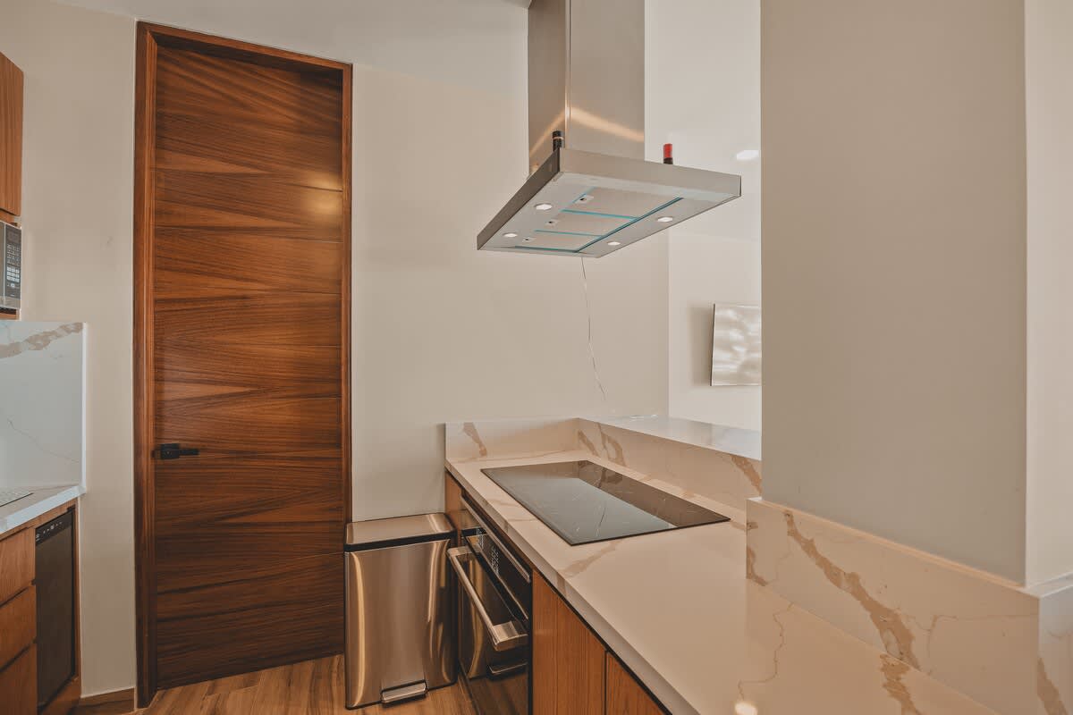 Modern kitchen corner showcasing an induction cooktop, built-in oven, and a stainless steel trash can, complemented by warm wooden cabinetry and white marble countertops.