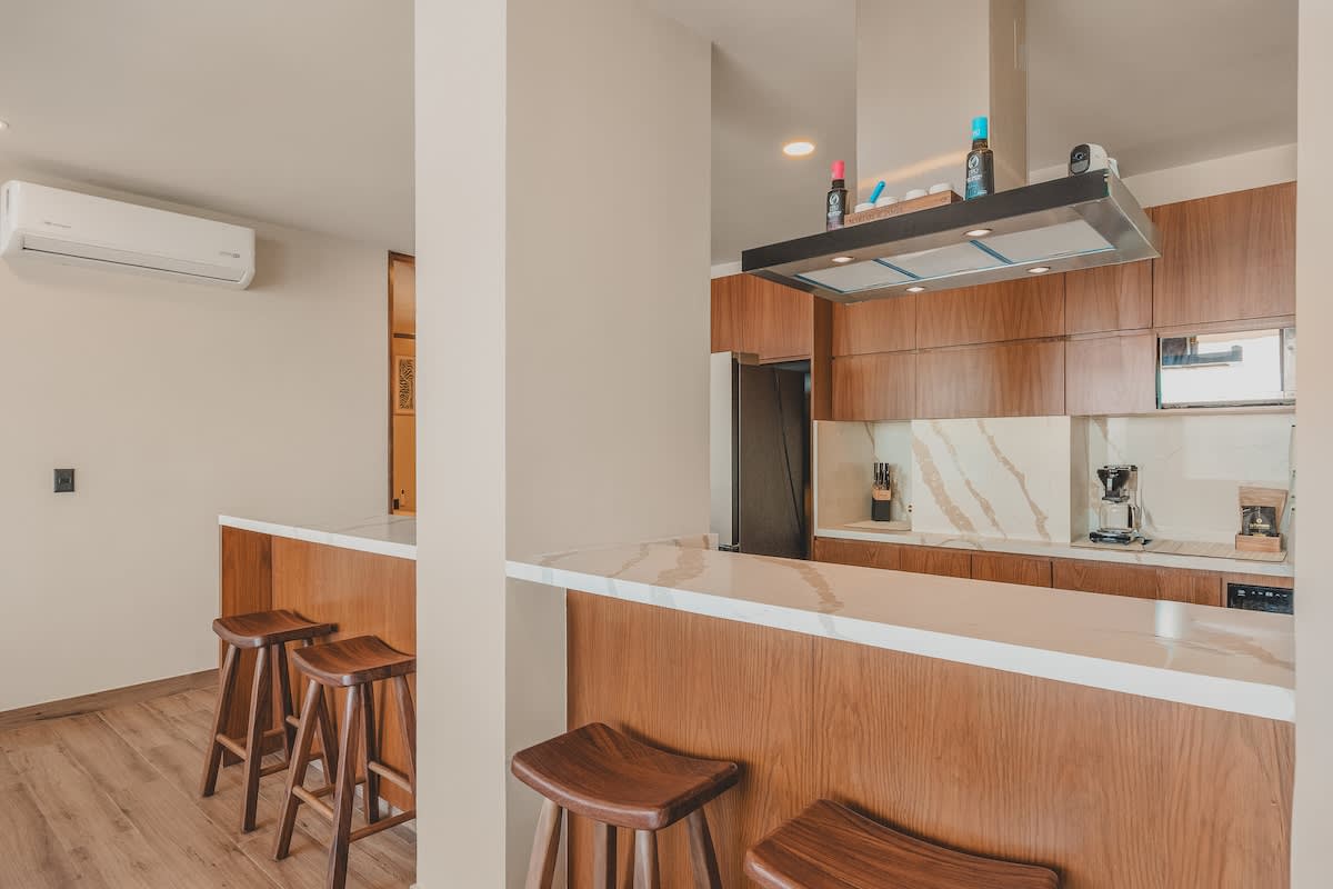 View of the modern kitchen with warm wood cabinets, white marble countertops, and a stainless steel range hood over an induction cooktop, featuring an air conditioning unit on the adjacent wall.