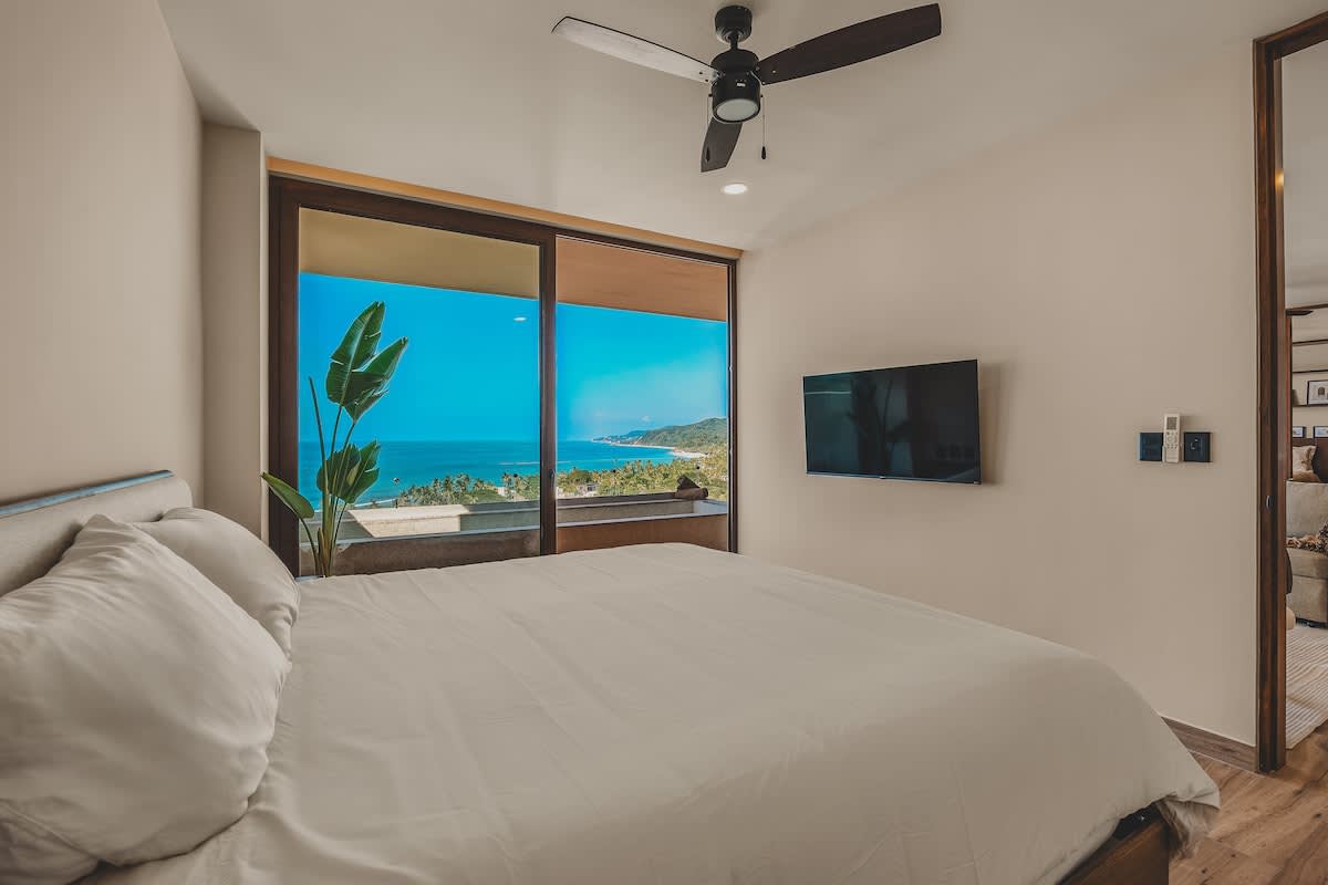 Bright bedroom featuring a queen-sized bed with white linens, a wall-mounted TV, and a dark ceiling fan, all set against a stunning ocean and coastline view through expansive sliding glass doors.