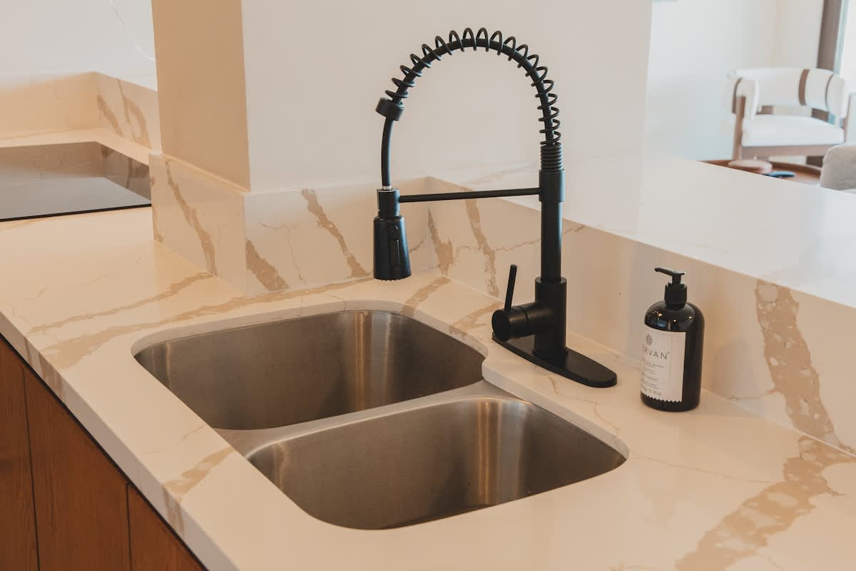 A modern kitchen features a double stainless steel sink with a stylish black pull-down faucet, set into a light-colored quartz countertop with prominent brown veining. A black soap dispenser sits beside the sink, ready for use.