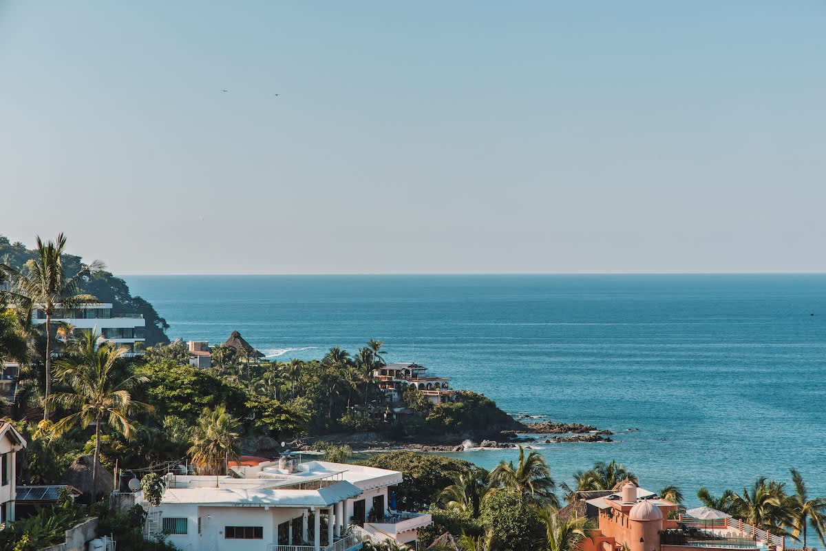Expansive ocean view from a high vantage point, showing the deep blue sea meeting a rocky coastline dotted with white buildings and lush green vegetation under a clear sky.