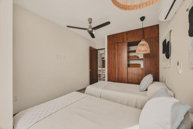 A bright bedroom features two twin beds with light bedding, a large built-in wooden wardrobe with open shelving, and a ceiling fan. A doorway leads to another area, possibly a hallway or bathroom.