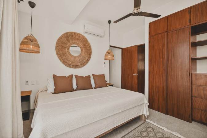 Serene bedroom with a large bed, earthy brown pillows, a prominent round woven mirror above the headboard, and a built-in wooden wardrobe. A ceiling fan and a woven pendant light enhance the natural aesthetic.