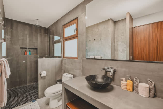 Contemporary bathroom featuring dark gray concrete walls and a matching vanity with a black vessel sink. A walk-in shower with black mosaic tiles and a toilet are visible, with a large mirror above the sink.