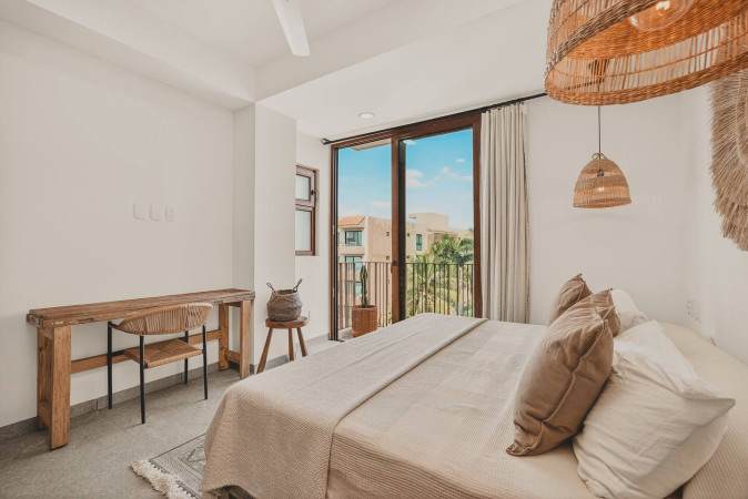 Bright bedroom featuring a large bed, a rustic wooden desk with a chair, and a sliding glass door leading to a balcony with views of buildings and lush greenery. Woven pendant lights add a natural touch.