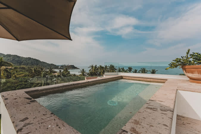 Rooftop swimming pool with a clear glass railing, offering breathtaking panoramic views of the ocean, distant coastline, and lush green mountains under a partly cloudy sky.