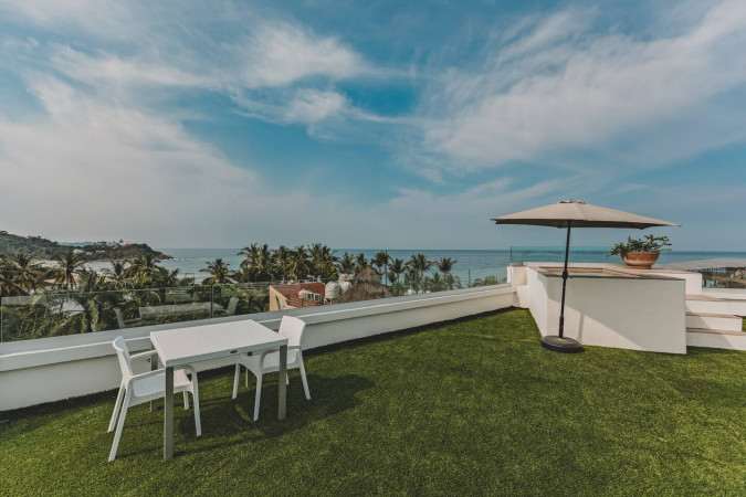 Rooftop deck with artificial grass, a white outdoor dining table with chairs, and a large umbrella, offering a stunning panoramic view of the ocean and palm trees along the coast.