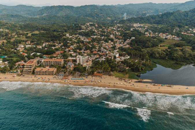 High aerial view of a vibrant coastal town featuring a long sandy beach with ocean waves, a river meeting the sea, and numerous buildings nestled among green foliage.