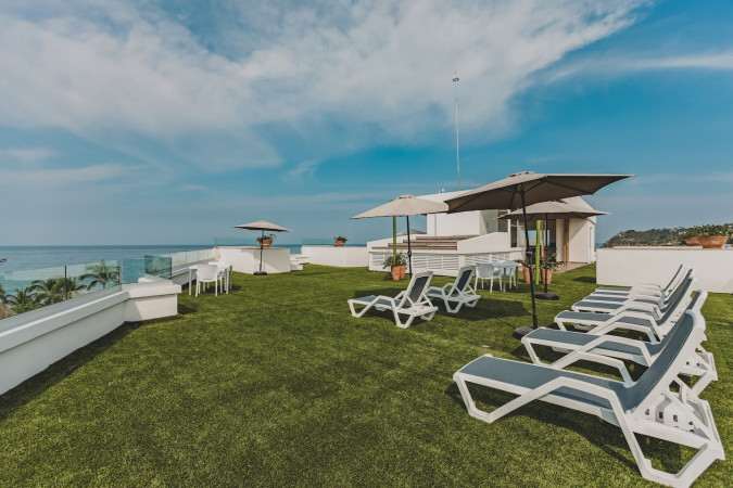 Wide shot of the rooftop deck featuring artificial turf, several lounge chairs, and two large umbrellas, with a clear view of the blue ocean and a distant coastline under a bright sky.