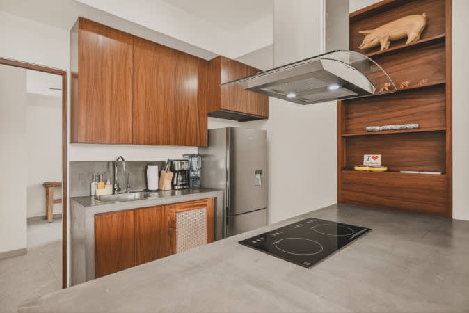 Well-equipped modern kitchen with a sleek concrete countertop, stainless steel sink, ample wooden upper and lower cabinets, and a full-size stainless steel refrigerator. An induction cooktop is integrated into the island.