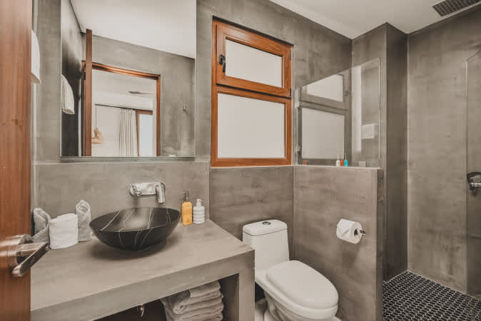 This contemporary bathroom features concrete-textured walls, a black vessel sink on a concrete vanity, a white toilet, and a shower area with dark hexagonal floor tiles. A wooden-framed window adds natural light.