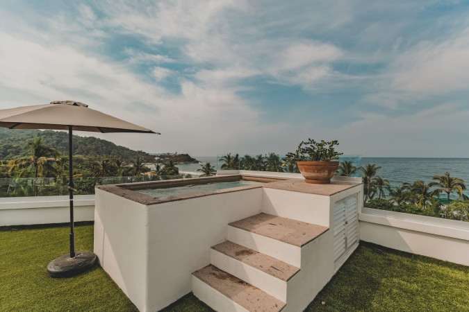Rooftop deck featuring a built-in hot tub with steps, artificial turf, and a large beige umbrella, offering panoramic views of the ocean, sandy beach, and lush green hills with palm trees under a partly cloudy sky.