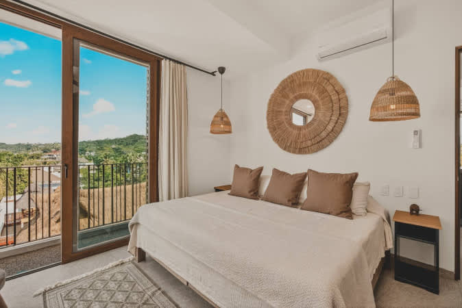 Bright bedroom with a large bed, neutral bedding, and brown throw pillows. A large circular woven mirror and two woven pendant lights adorn the wall, with a balcony offering a scenic view through sliding glass doors.