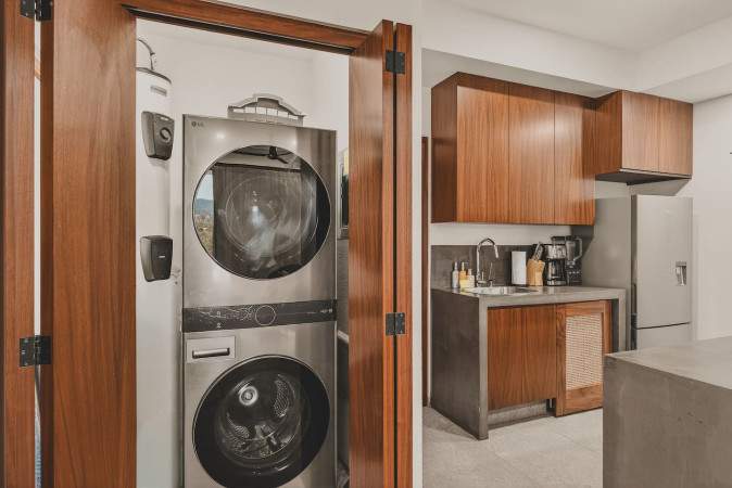 A compact laundry area with a stacked silver washer and dryer unit is visible through open wooden bi-fold doors, adjacent to a modern kitchen with dark wood cabinetry and concrete countertops.
