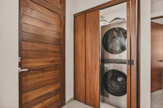 A compact laundry closet features a stacked silver washer and dryer unit, neatly tucked behind warm-toned wooden bi-fold doors, adjacent to another solid wooden door.