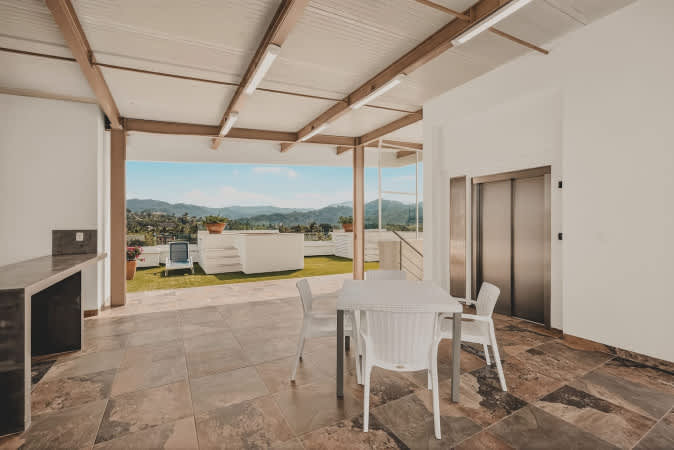 Rooftop deck area with an elevator and a white dining table set, offering expansive views of rolling green hills and mountains in the distance, with a glimpse of a pool.