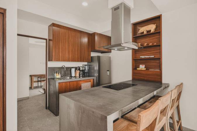 Modern kitchen with dark wood cabinetry, a concrete island with an induction cooktop, and three wooden bar stools. A stainless steel range hood and refrigerator are visible, along with open shelving.