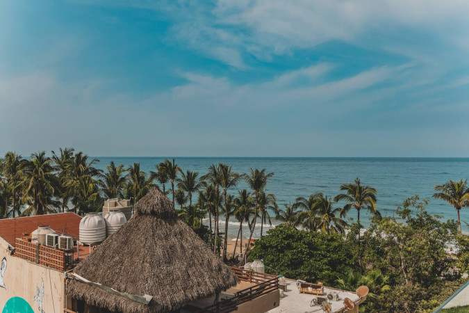 Scenic view of the ocean and a sandy beach lined with numerous palm trees, seen from a high vantage point, with traditional thatched roofs visible in the foreground.