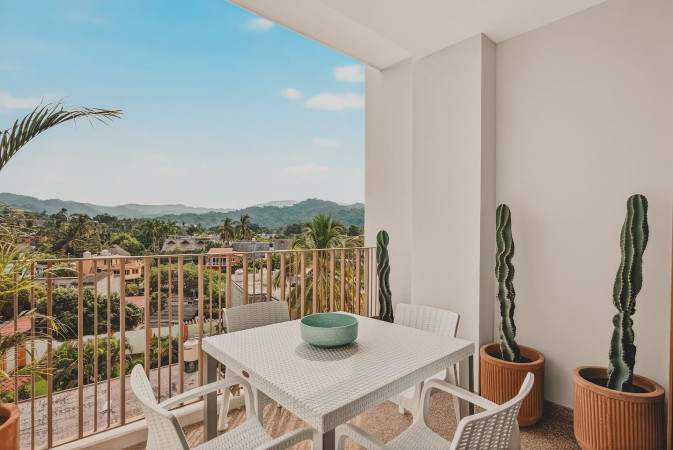 Balcony with a white outdoor dining set and potted cacti, overlooking a vibrant town and distant hills under a bright blue sky. The metal railing provides an open view.