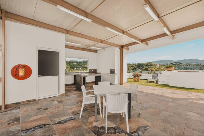 Covered outdoor kitchen and dining area with a patterned tiled floor, a white dining table with chairs, a built-in counter with a sink, and a fire extinguisher, overlooking distant mountains.
