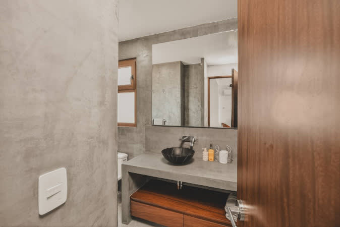 View into a modern bathroom showcasing textured concrete walls and a matching vanity with a sleek black vessel sink, illuminated by a large mirror. A warm wooden door frame is visible on the right.