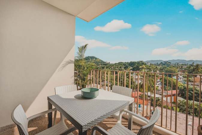 Balcony with a white wicker dining table and chairs, offering a panoramic view of a lush green landscape with distant mountains and a clear blue sky. A metal railing encloses the space.
