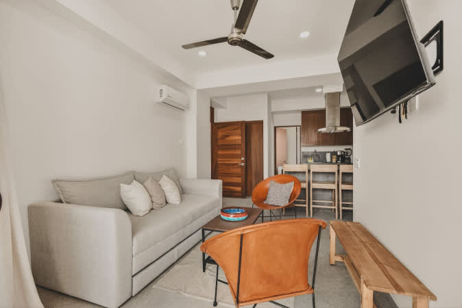 Modern open-concept living area with a light gray sofa, two orange leather lounge chairs, and a wooden coffee table. A ceiling fan and wall-mounted TV are visible, with a compact kitchen in the background.