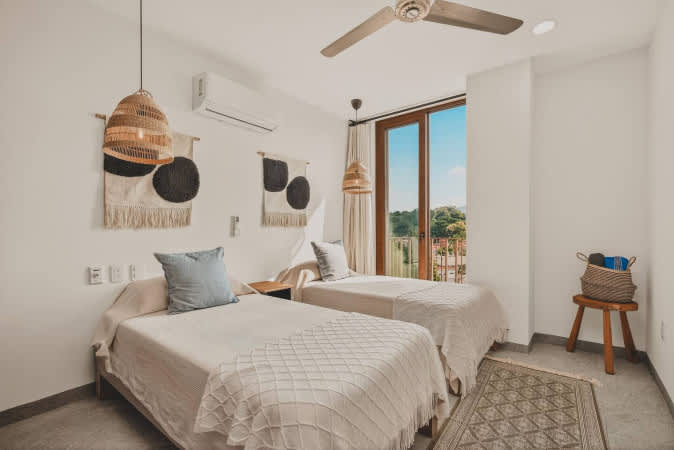 This bright bedroom offers two twin beds with light-colored bedding, a large glass door leading to a balcony with scenic views, and a ceiling fan for comfort. Woven pendant lights and wall art add a touch of bohemian style.