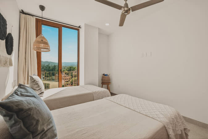 A serene bedroom features two twin beds with light bedding and a large glass door opening to a balcony with lush green views. A ceiling fan provides air circulation, and a woven basket sits on a small wooden stool.