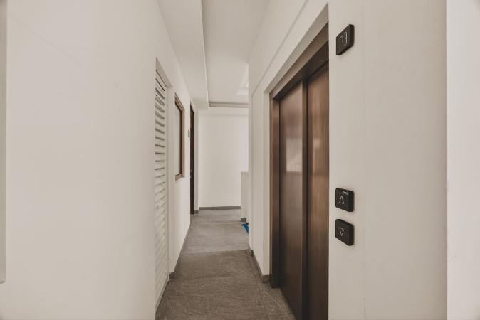 Building hallway with an elevator featuring a wooden door and control panel. White walls and a light gray floor extend down the corridor.