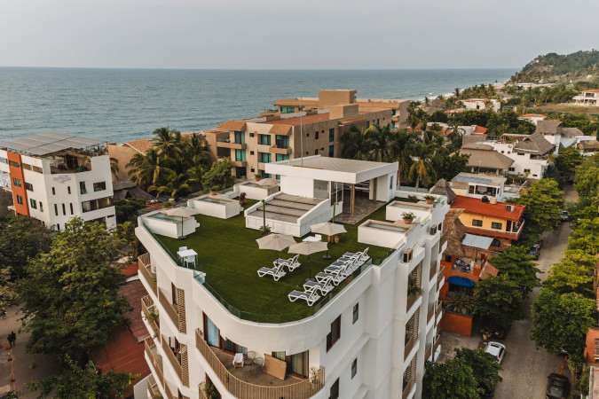 Aerial view showcasing the white apartment building's rooftop with artificial grass, numerous lounge chairs, and umbrellas, offering a glimpse of the ocean and coastline in the distance.