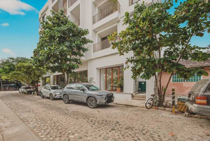White multi-story building with balconies and large windows, situated on a cobblestone street with parked cars and lush green trees. The architecture is modern and clean.