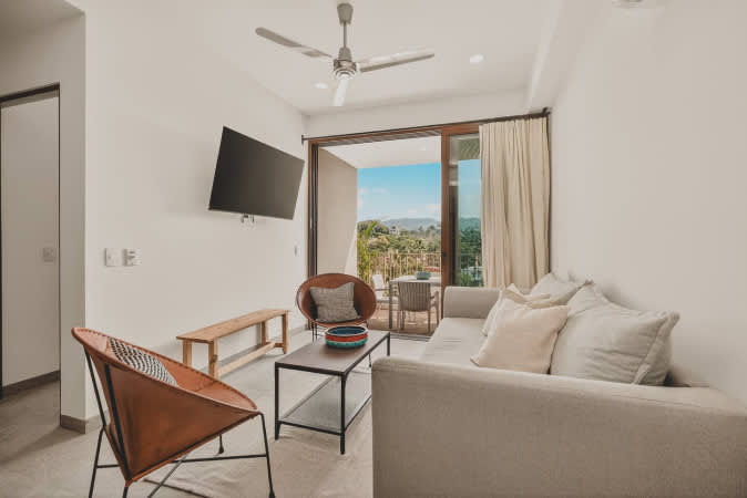 Bright and airy living room furnished with a comfortable light grey sofa, two stylish orange accent chairs, a minimalist coffee table, and a wall-mounted TV. A large sliding glass door opens to a balcony with scenic views.