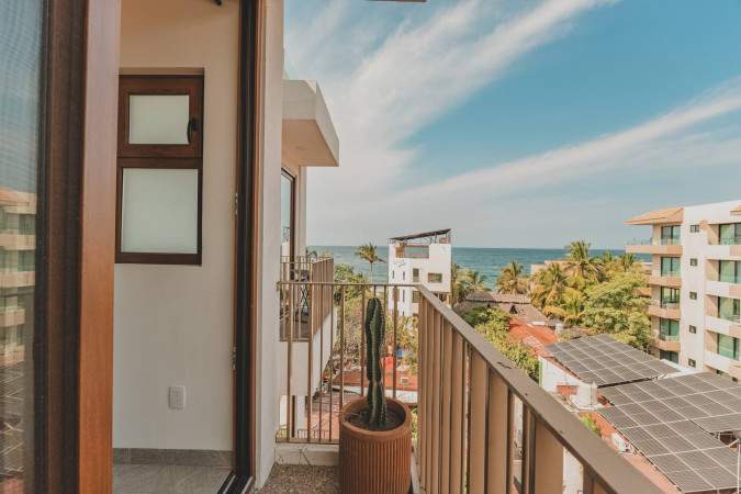 A private balcony offers a stunning view of the ocean and surrounding buildings, featuring a metal railing and a potted cactus. The wooden door frame adds a natural touch to the outdoor space.